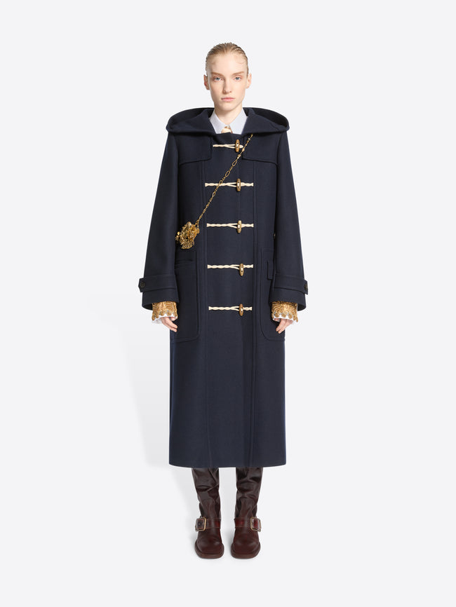 A model stands against a light gray background wearing a long navy duffle coat with toggle closures, a large dark hood and gold chain strap bag, paired with brown boots.
