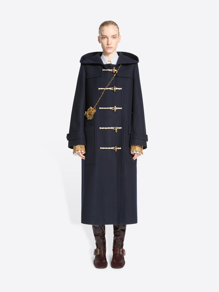 A model stands against a light gray background wearing a long navy duffle coat with toggle closures, a large dark hood and gold chain strap bag, paired with brown boots.