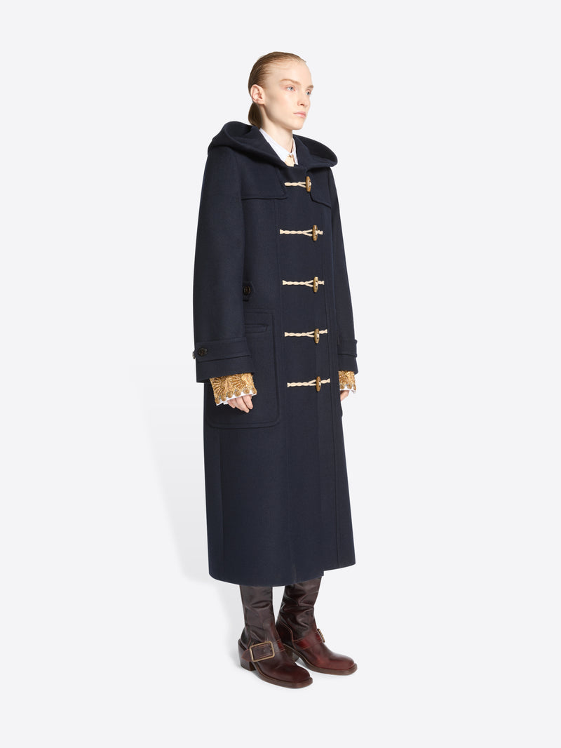 A model stands against a light gray background wearing a long navy duffle coat with toggle closures, a large dark hood, paired with brown boots.