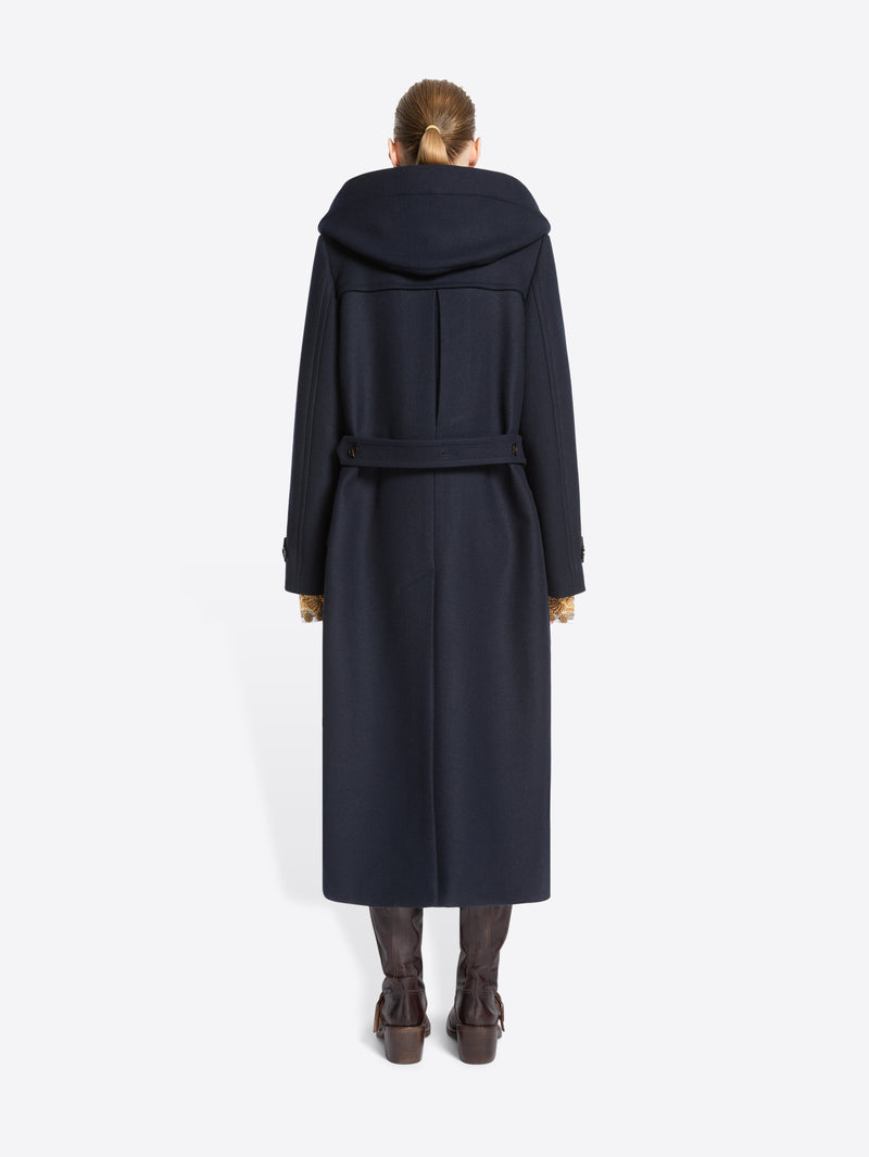Back view of a model standing against a light gray background wearing a long navy duffle coat with large dark hood, paired with brown boots.