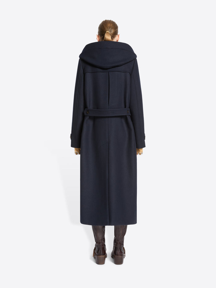Back view of a model standing against a light gray background wearing a long navy duffle coat with large dark hood, paired with brown boots.