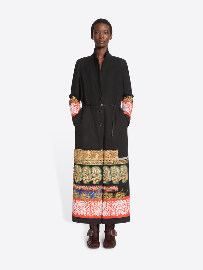 A model stands against a light gray background wearing a long black wrap-style coat with ornate multicolor patterned bands around the hem and cuffs, paired with dark brown boots.