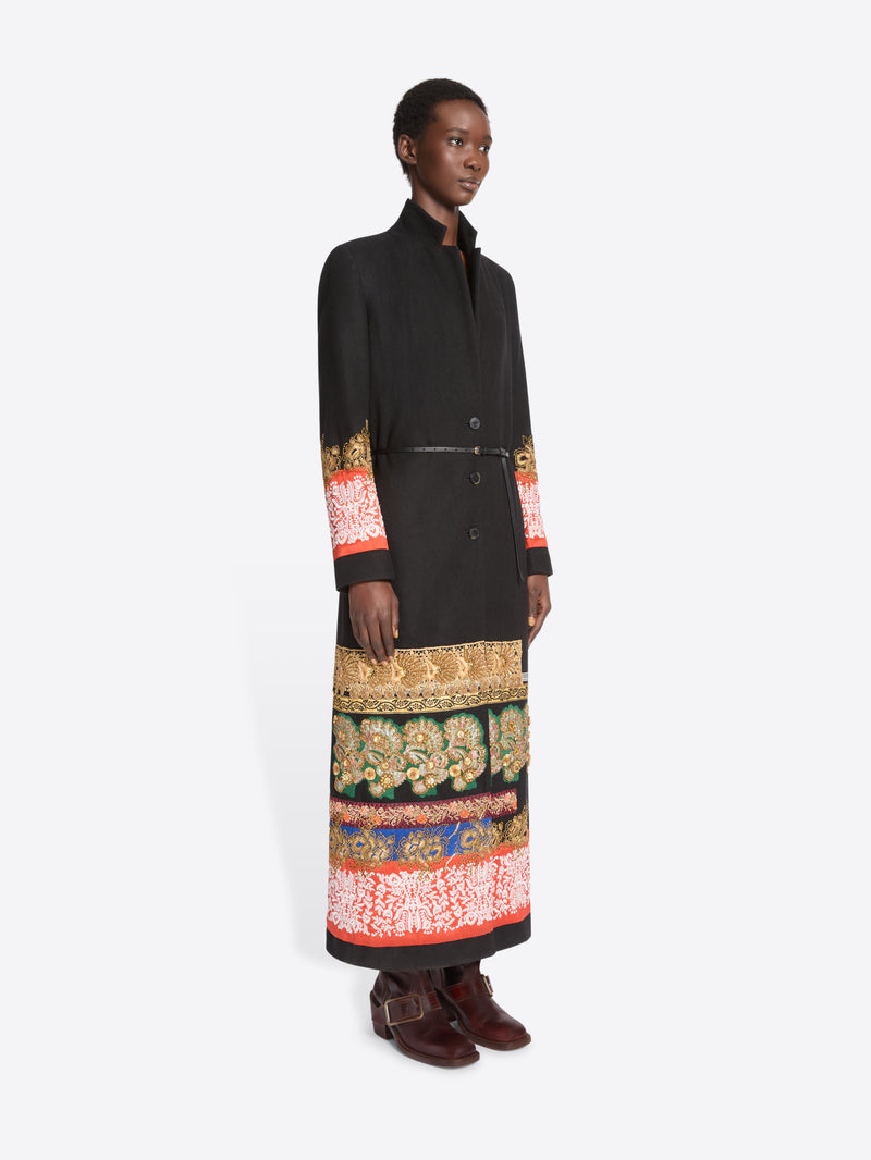 A model stands against a light gray background wearing a long black wrap-style coat with ornate multicolor patterned bands around the hem and cuffs, paired with dark brown boots.
