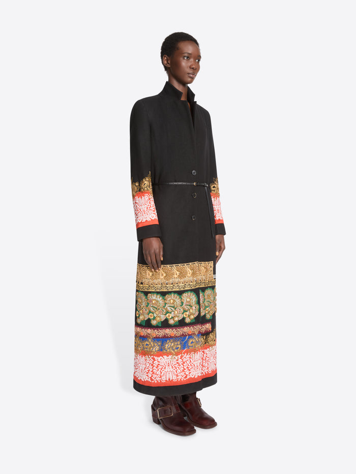 A model stands against a light gray background wearing a long black wrap-style coat with ornate multicolor patterned bands around the hem and cuffs, paired with dark brown boots.