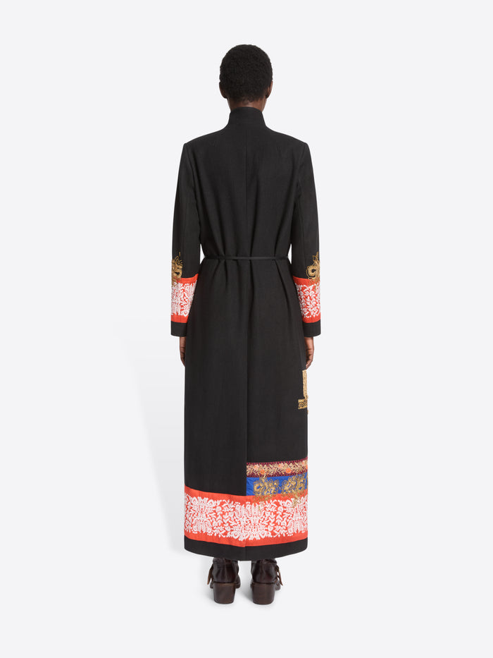 Back view of a model standing against a light gray background wearing a long black wrap-style coat with ornate multicolor patterned bands around the hem and cuffs, paired with dark brown boots.