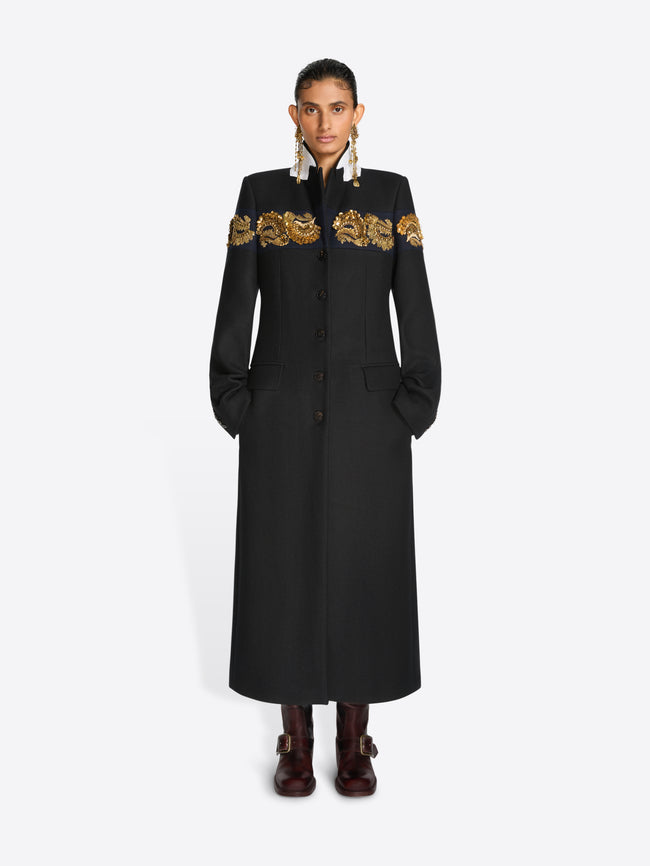 A model stands against a light gray background wearing a long black coat with ornate gold embroidery across the shoulders and upper chest, paired with dark brown boots and large dangling earrings.