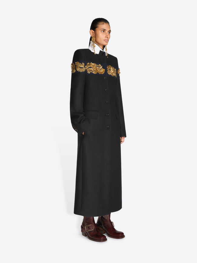 A model stands against a light gray background wearing a long black coat with ornate gold embroidery across the shoulders and upper chest, paired with dark brown boots and large dangling earrings.
