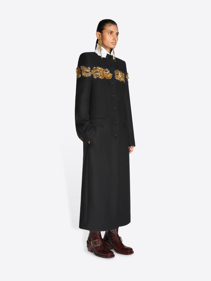 A model stands against a light gray background wearing a long black coat with ornate gold embroidery across the shoulders and upper chest, paired with dark brown boots and large dangling earrings.