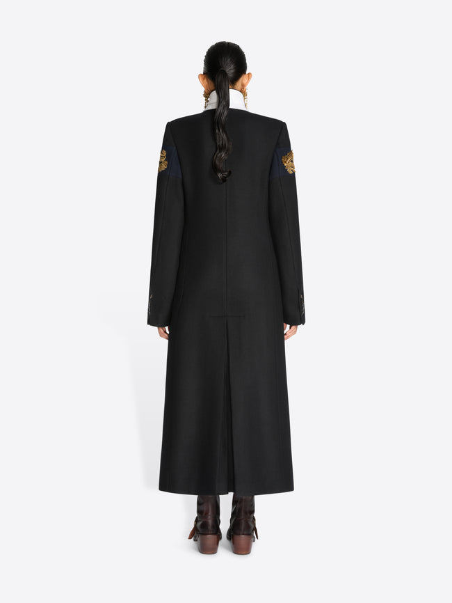 Back view of a model wearing a long black coat with embroidered details and brown leather boots, set against a plain light gray background.