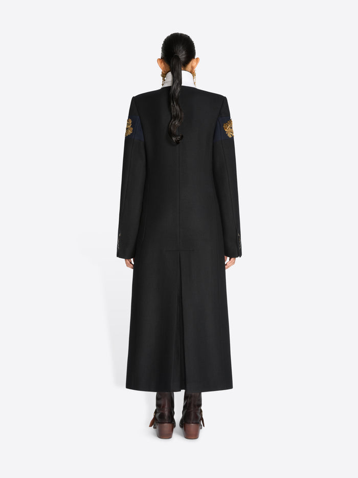 Back view of a model wearing a long black coat with embroidered details and brown leather boots, set against a plain light gray background.