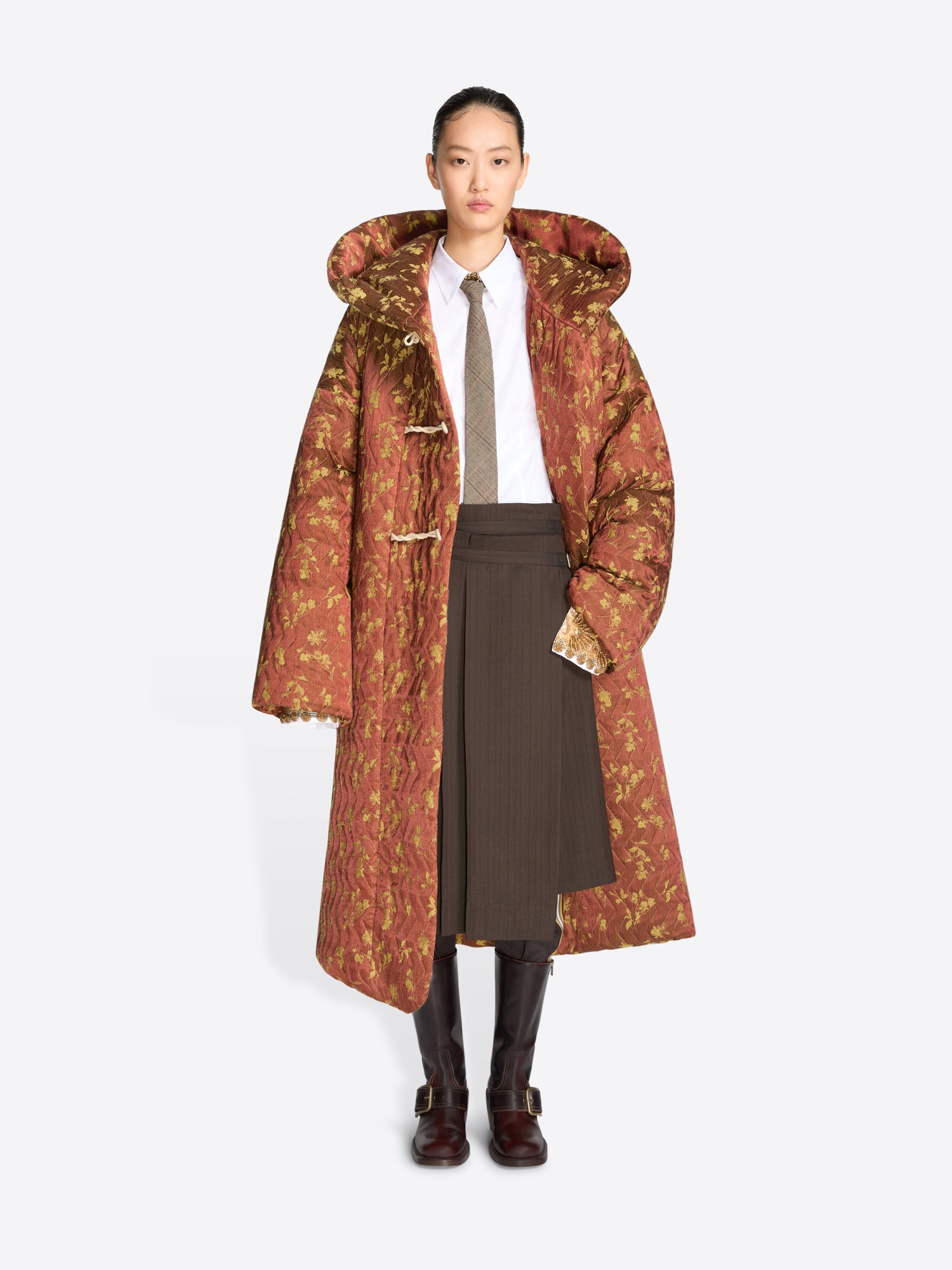 A model stands against a light gray background wearing a long, oversized hooded coat with a rust-brown and olive floral pattern, layered over a white shirt, taupe tie, pleated brown skirt, and dark brown boots.