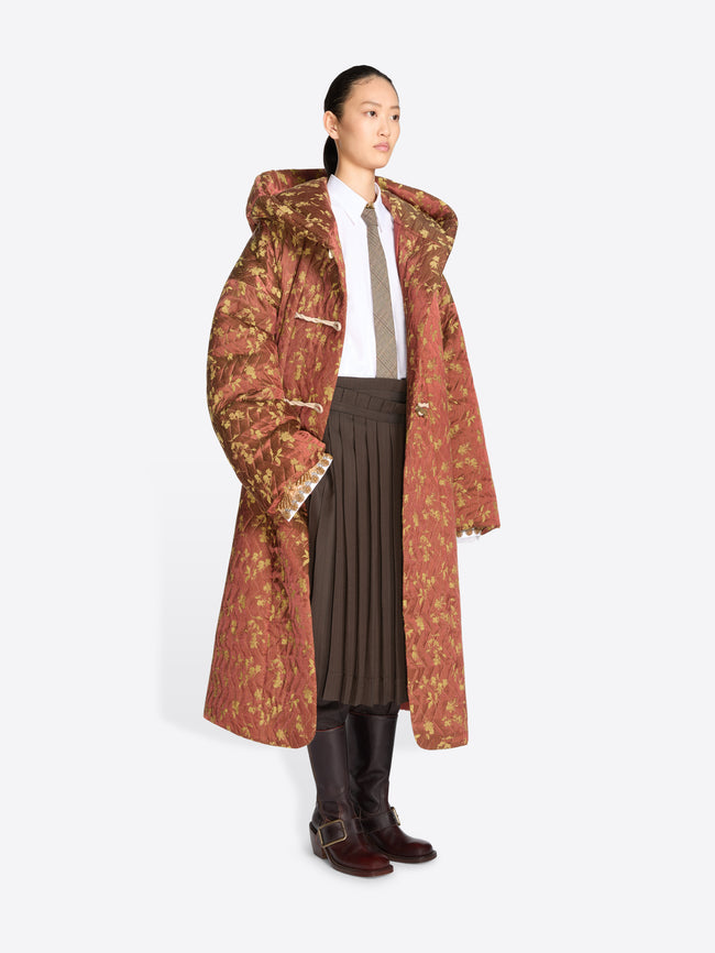 A model stands against a light gray background wearing a long, oversized hooded coat with a rust-brown and olive floral pattern, layered over a white shirt, taupe tie, pleated brown skirt, and dark brown boots.