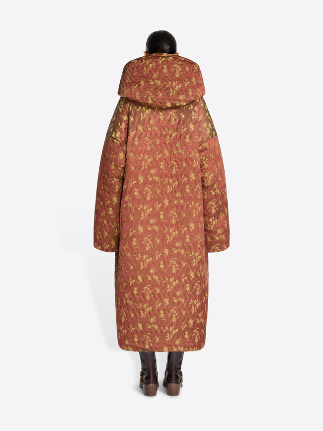 Back view of a model standing against a light gray background wearing a long, oversized hooded coat with a rust-brown and olive floral pattern and dark brown boots.