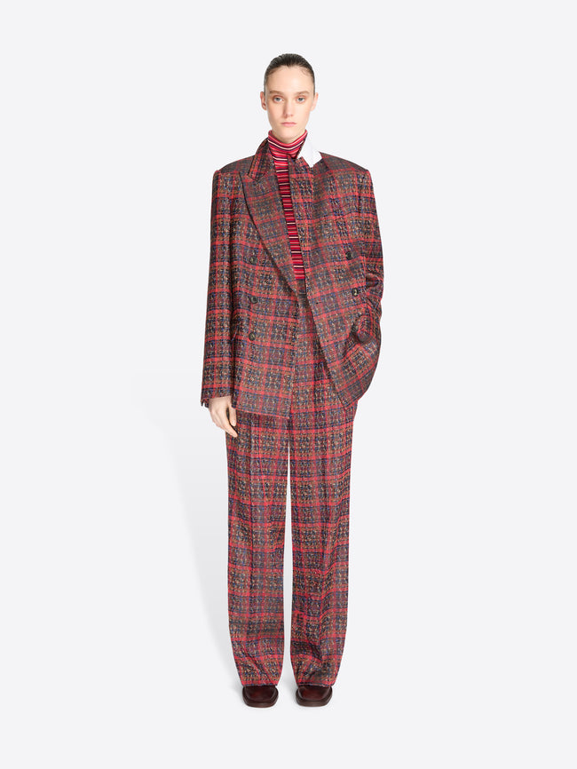 A model stands against a light gray background wearing a matching red and black plaid suit, featuring a double-breasted blazer and wide-leg pants, styled with a red striped turtleneck.