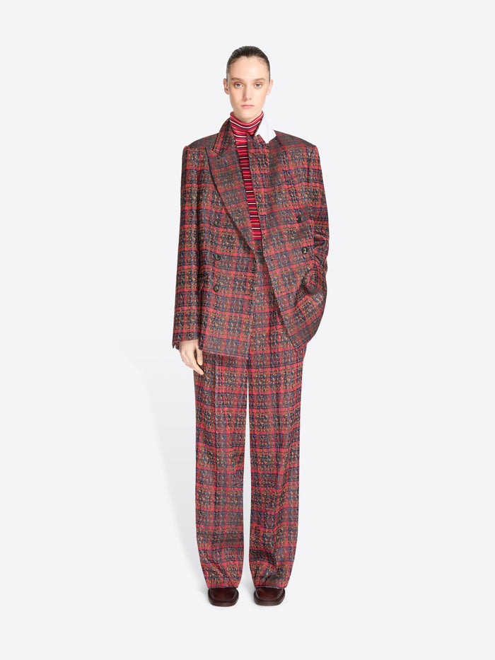 A model stands against a light gray background wearing a matching red and black plaid suit, featuring a double-breasted blazer and wide-leg pants, styled with a red striped turtleneck.