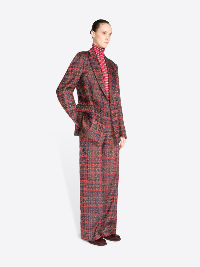 A model stands against a light gray background wearing a matching red and black plaid suit, featuring a double-breasted blazer and wide-leg pants, styled with a red striped turtleneck.