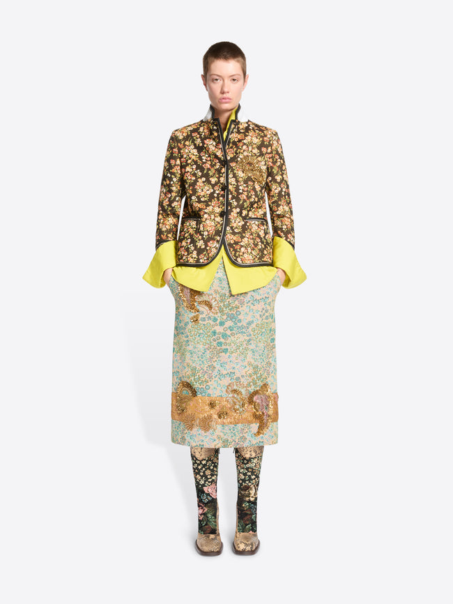 A model stands against a light gray background wearing a full-length patterned outfit. The look features a floral patterned cropped jacket featuring the bright yellow flared shirt, a matching patterned skirt and patterned tights.