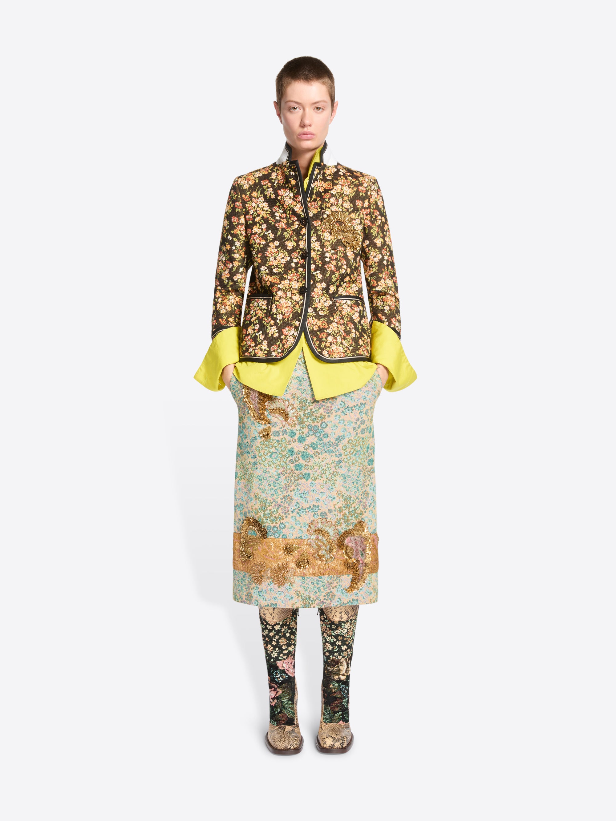 A model stands against a light gray background wearing a full-length patterned outfit. The look features a floral patterned cropped jacket featuring the bright yellow flared shirt, a matching patterned skirt and patterned tights.
