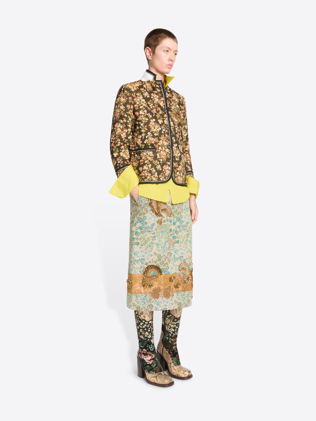 A model stands against a light gray background wearing a full-length patterned outfit. The look features a floral patterned cropped jacket featuring the bright yellow flared shirt, a matching patterned skirt and patterned tights.