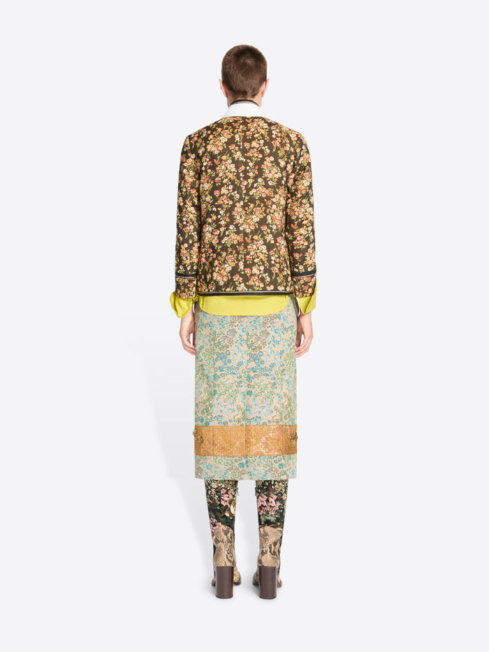 Back view of a modelmwearing a long-sleeved floral jacket, a patterned skirt with a gold band, matching patterned tights, and heels against a light gray background.