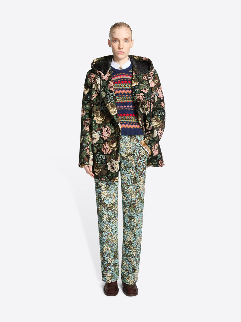 A model stands against a light gray background wearing a dark floral-patterned coat over a multicolor sweater, matching floral pants and dark brown shoes.