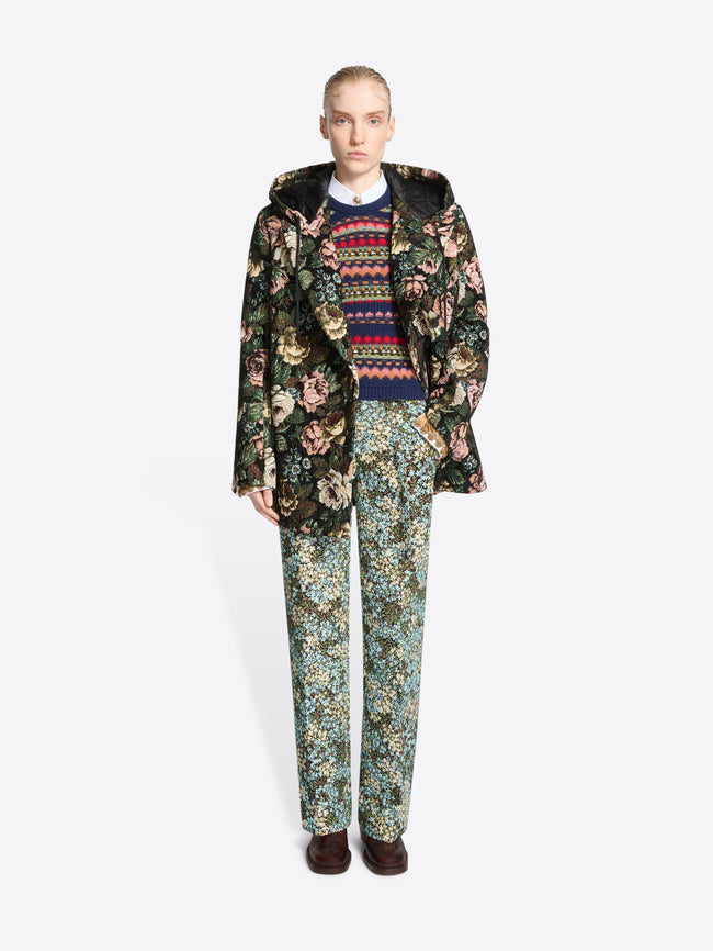 A model stands against a light gray background wearing a dark floral-patterned coat over a multicolor sweater, matching floral pants and dark brown shoes.