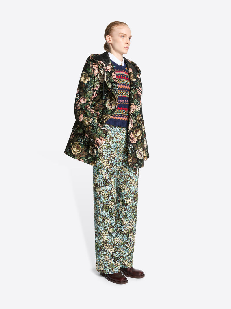 A model stands against a light gray background wearing a dark floral-patterned coat over a multicolor sweater, matching floral pants and dark brown shoes.