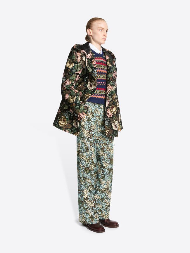 A model stands against a light gray background wearing a dark floral-patterned coat over a multicolor sweater, matching floral pants and dark brown shoes.
