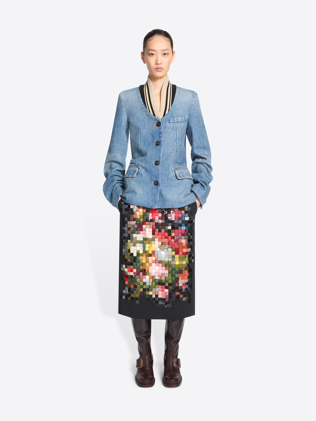 A model stands against a light gray background wearing a light blue denim jacket with black and beige trim, paired with a black midi skirt featuring a colorful pixelated floral pattern, and dark brown boots.