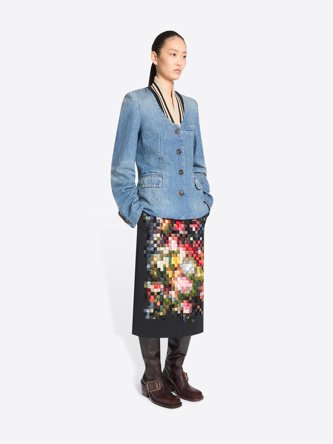 A model stands against a light gray background wearing a light blue denim jacket with black and beige trim, paired with a black midi skirt featuring a colorful pixelated floral pattern, and dark brown boots.