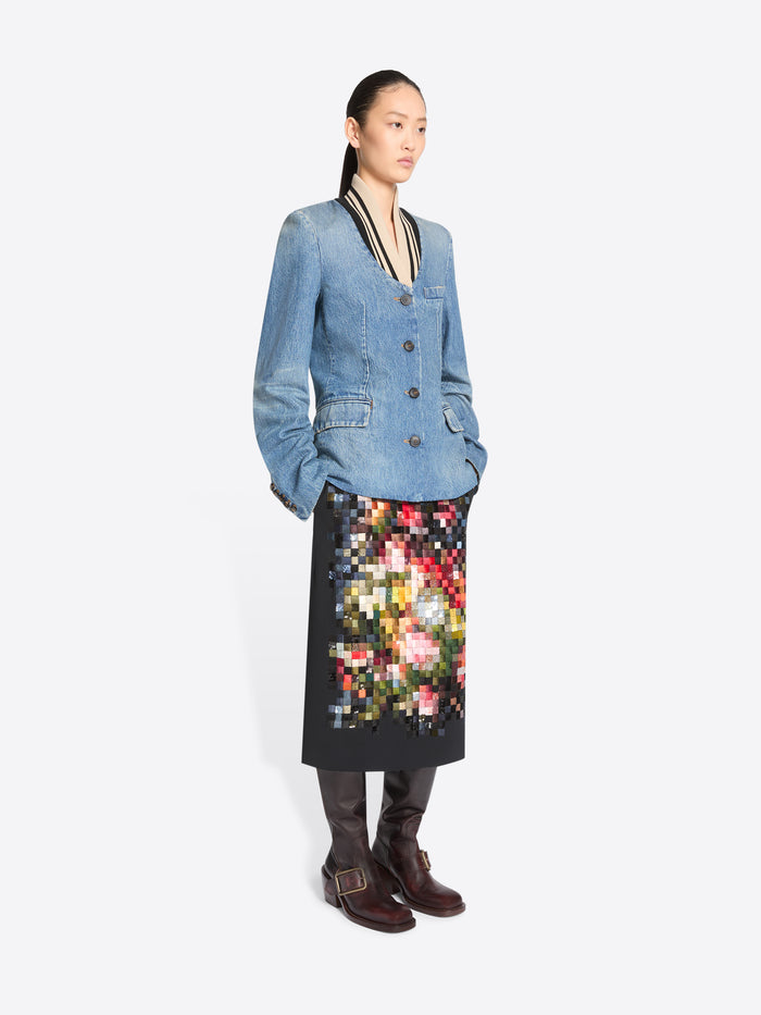 A model stands against a light gray background wearing a light blue denim jacket with black and beige trim, paired with a black midi skirt featuring a colorful pixelated floral pattern, and dark brown boots.