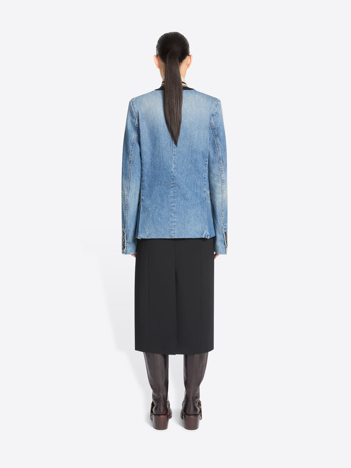 Back view of a model standing against a light gray background wearing a light blue denim jacket with a black midi skirt and dark brown boots.