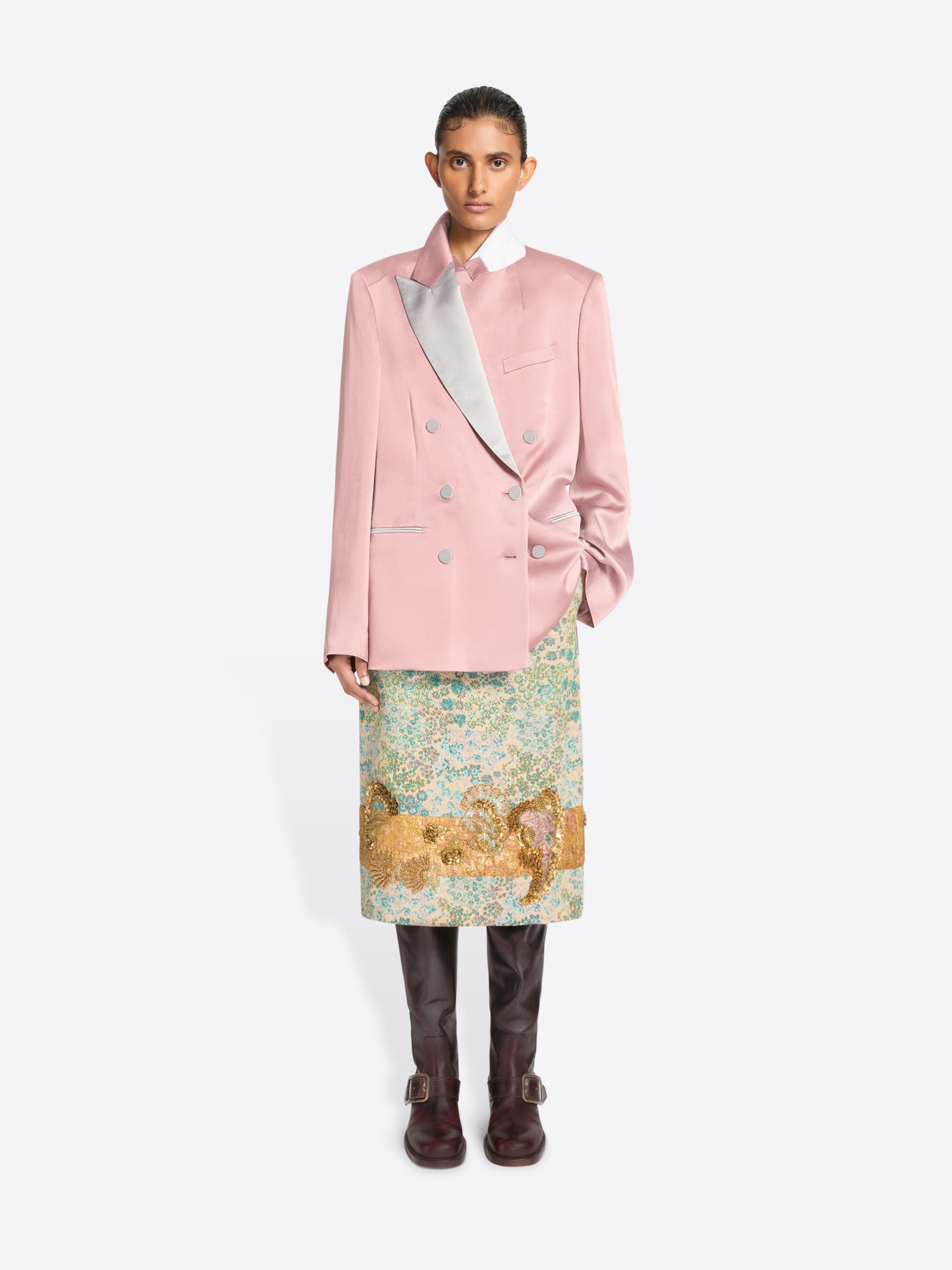 A model stands against a light gray background wearing a pink double-breasted blazer with a contrasting light gray lapel, a patterned green and gold skirt and dark brown boots.