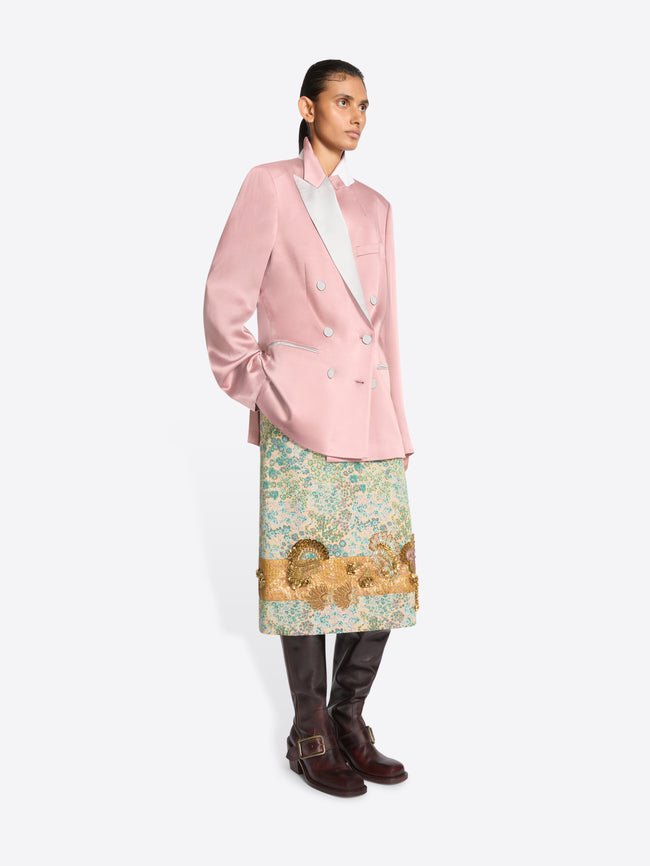 A model stands against a light gray background wearing a pink double-breasted blazer with a contrasting light gray lapel, a patterned green and gold skirt and dark brown boots.