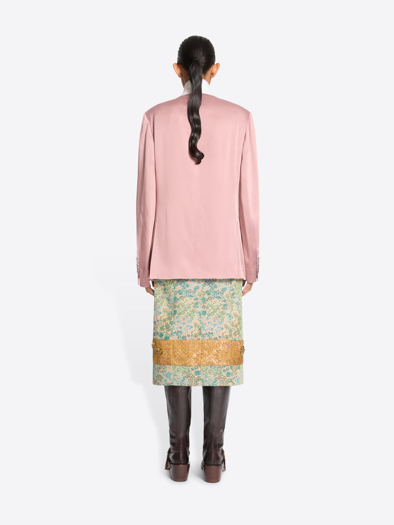 Back view of a model standing against a light gray background wearing a pink double-breasted blazer, a patterned green and gold skirt and dark brown boots.
