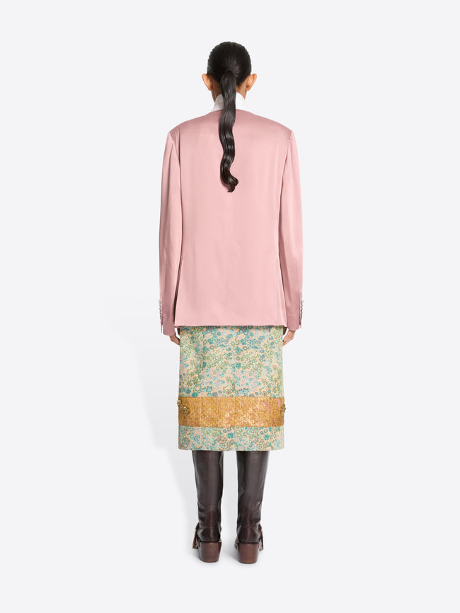 Back view of a model standing against a light gray background wearing a pink double-breasted blazer, a patterned green and gold skirt and dark brown boots.
