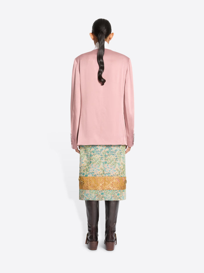 Back view of a model standing against a light gray background wearing a pink double-breasted blazer, a patterned green and gold skirt and dark brown boots.