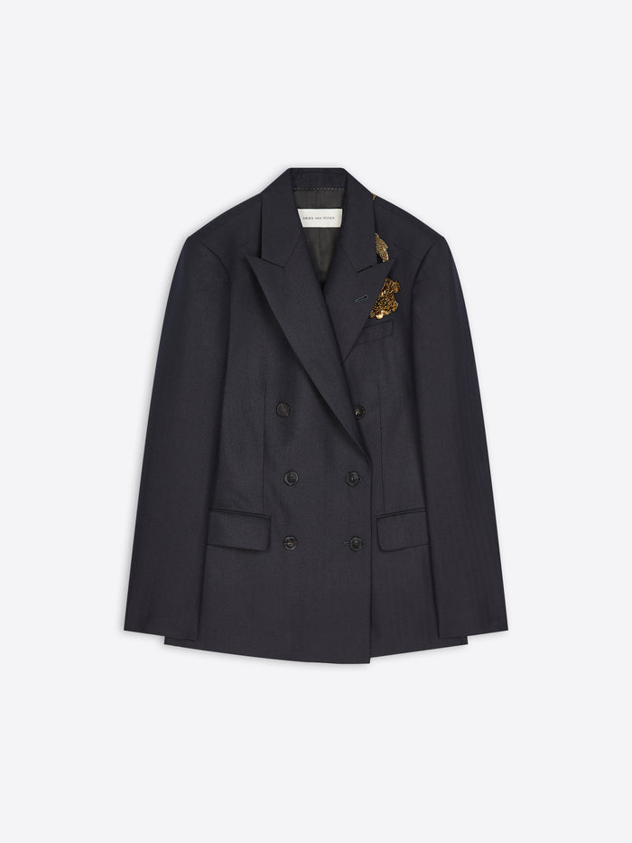 A dark double-breasted blazer with a small gold embroidered detail on the lapel, displayed flat against a light gray background.