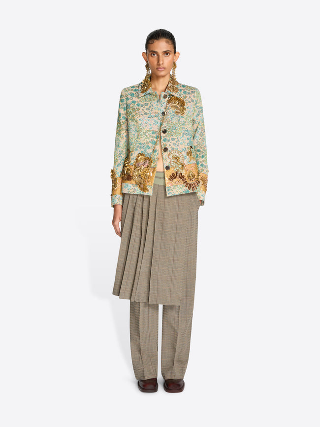 A model stands against a light gray background wearing a light green and gold patterned jacket over a beige pleated skirt and matching wide-leg trousers.