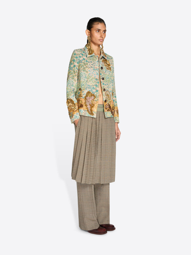 A model stands against a light gray background wearing a light green and gold patterned jacket over a beige pleated skirt and matching wide-leg trousers.