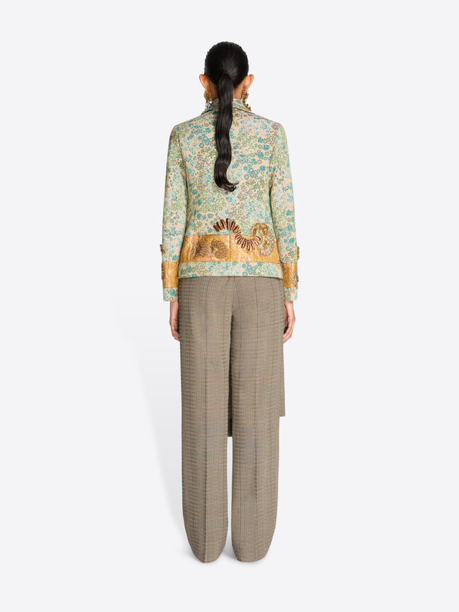 Back view of a model standing against a light gray background wearing a light green and gold patterned jacket over a beige pleated skirt and matching wide-leg trousers.