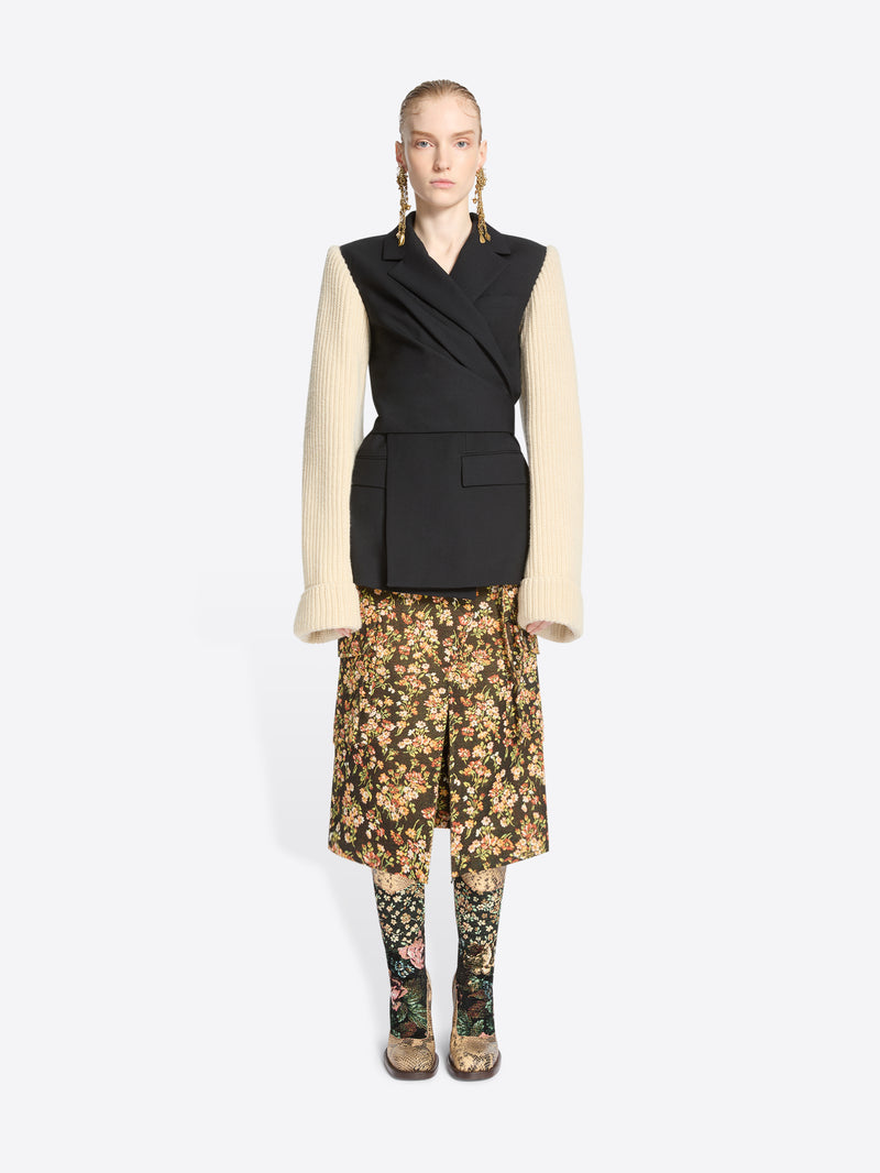 A model stands against a light gray background wearing a black wrap-style blazer with a black body and contrasting cream ribbed knit sleeves, paired with a floral midi skirt and matching floral tights.