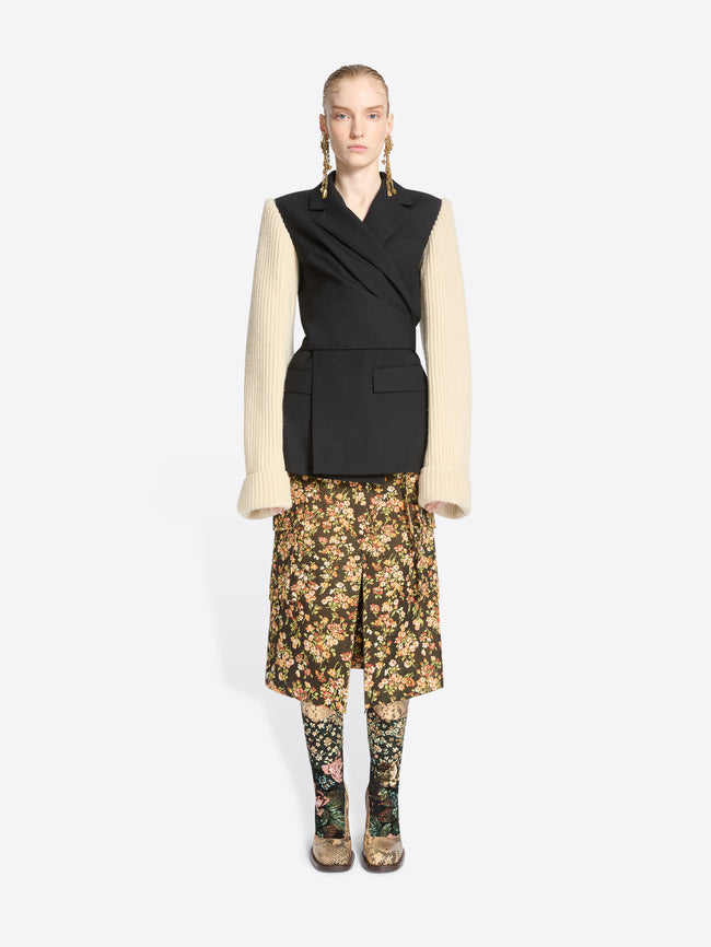 A model stands against a light gray background wearing a black wrap-style blazer with a black body and contrasting cream ribbed knit sleeves, paired with a floral midi skirt and matching floral tights.