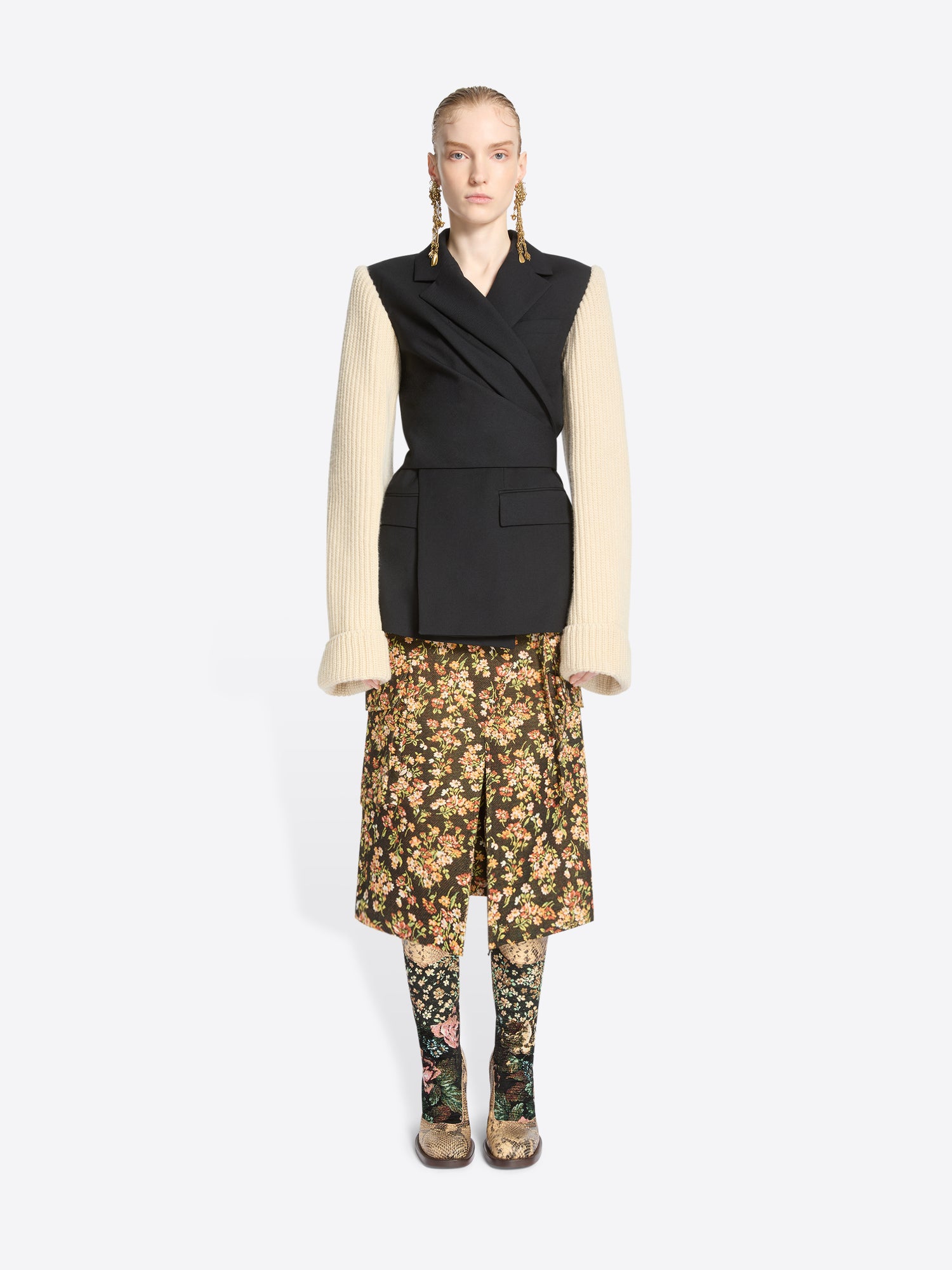A model stands against a light gray background wearing a black wrap-style blazer with a black body and contrasting cream ribbed knit sleeves, paired with a floral midi skirt and matching floral tights.