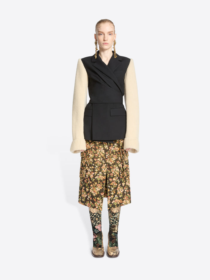 A model stands against a light gray background wearing a black wrap-style blazer with a black body and contrasting cream ribbed knit sleeves, paired with a floral midi skirt and matching floral tights.