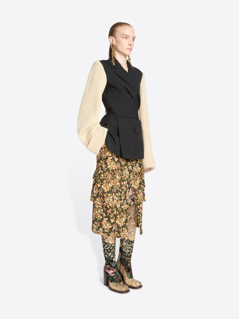 A model stands against a light gray background wearing a black wrap-style blazer with a black body and contrasting cream ribbed knit sleeves, paired with a floral midi skirt and matching floral tights.