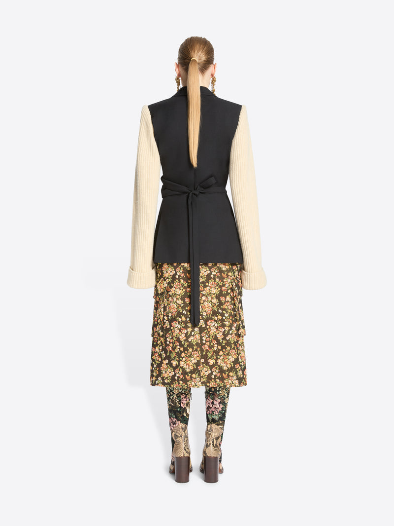 Back view of a model stands against a light gray background wearing a black wrap-style blazer with a black body and contrasting cream ribbed knit sleeves, paired with a floral midi skirt and matching floral tights.