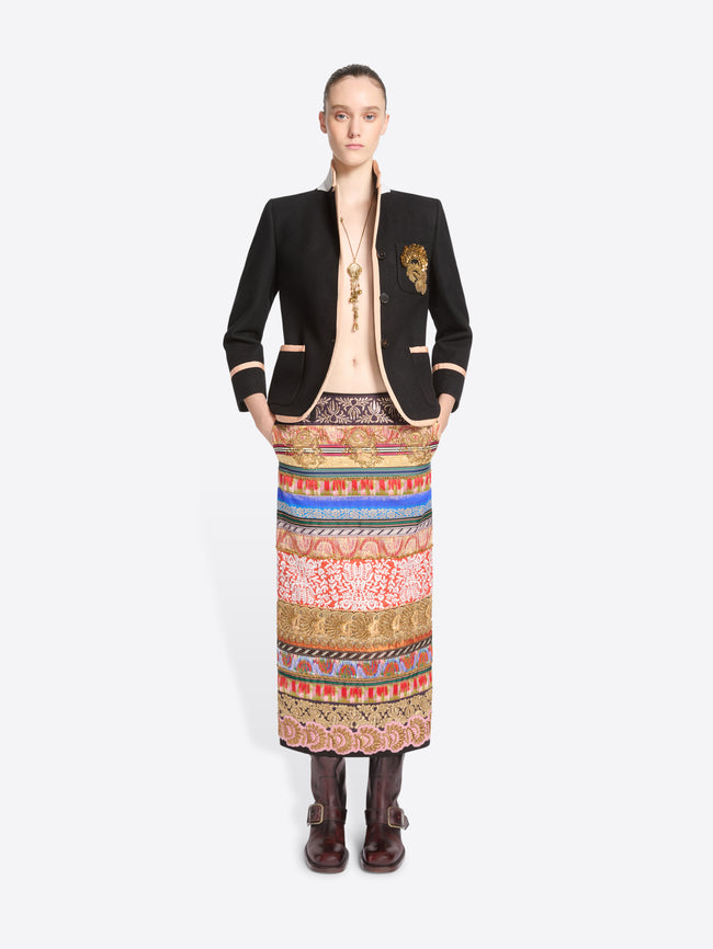 A model stands against a light gray background wearing a black blazer with pink trim and a long, wide, highly patterned skirt in red, blue, gold, and beige tones, paired with dark brown boots.