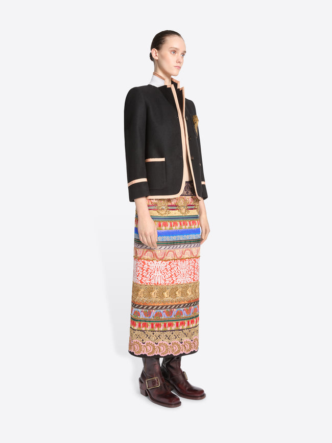 A model stands against a light gray background wearing a black blazer with pink trim and a long, wide, highly patterned skirt in red, blue, gold, and beige tones, paired with dark brown boots.