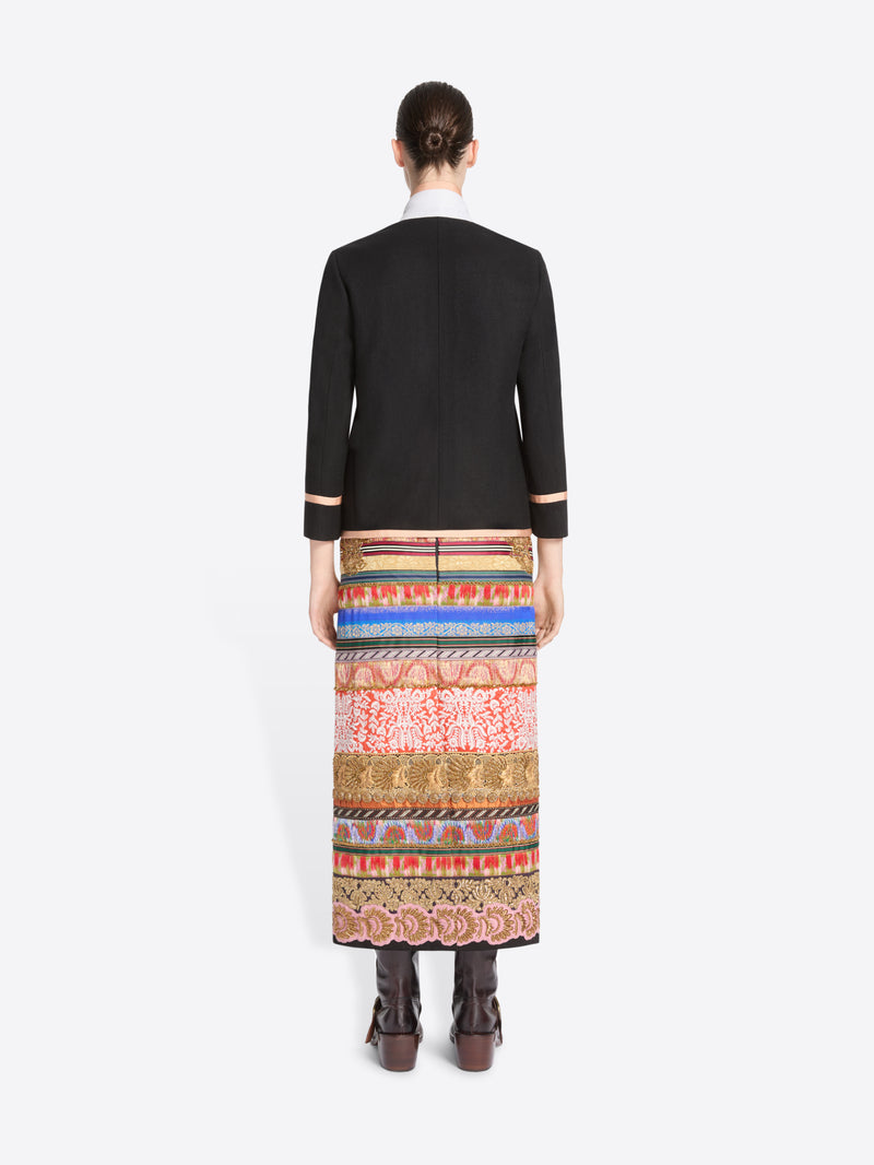 back view of a model stands against a light gray background wearing a black blazer with pink trim and a long, wide, highly patterned skirt in red, blue, gold, and beige tones, paired with dark brown boots.